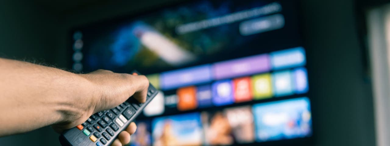 A man's hand holding a remote in front of a television screen with different streaming service icons